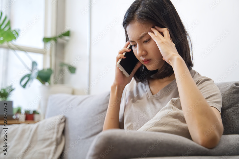 Upset/unhappy Asian girl talking on telephone is sitting on sofa at home. She feels uncomfortable and put the hand on temples. Social family, business and communication problem concept. Upset/unhappy Asian girl talking on telephone is sitting on sofa at home. She feels uncomfortable and put the hand on temples. Social family, business and communication problem concept.
