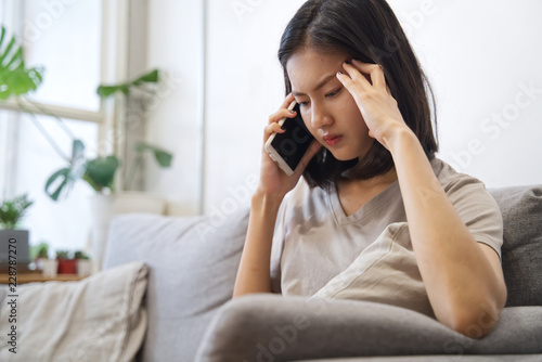 Upset/unhappy Asian girl talking on telephone is sitting on sofa at home. She feels uncomfortable and put the hand on temples. Social family, business and communication problem concept.