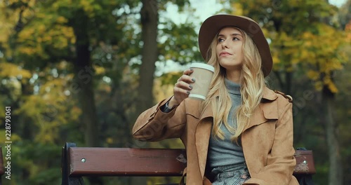 Caucasian blonde attractive and stylish young Caucasian woman sitting on the bench in a park, drinking coffee while waitingfor somebody and looking at the watch on her hand.