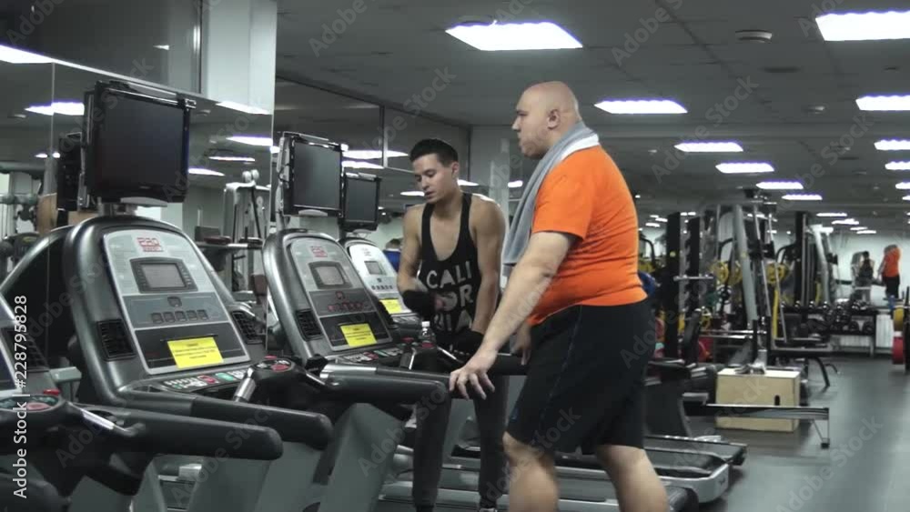 Fat man walking on the treadmill with a decisive look. Man's handsome young coach approves, praises, supports him. The view from the left side Fat man walking on the treadmill with a decisive look. Man's handsome young coach approves, praises, supports him. The view from the left side
