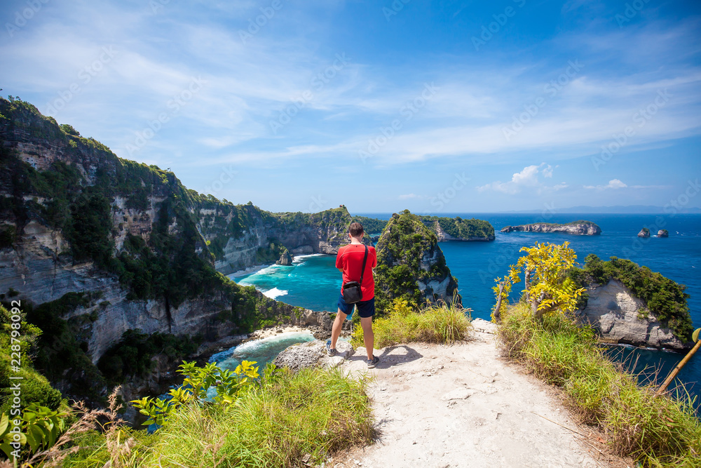 Man in red t-shirt standing on the rock and viewing Thousand islands Nusa Batumategan and Nusa Batupadasan from the Atuh Rija Lima shrine on Nusa Penida Island near Bali, Indonesia while sky is blue Man in red t-shirt standing on the rock and viewing Thousand islands Nusa Batumategan and Nusa Batupadasan from the Atuh Rija Lima shrine on Nusa Penida Island near Bali, Indonesia while sky is blue