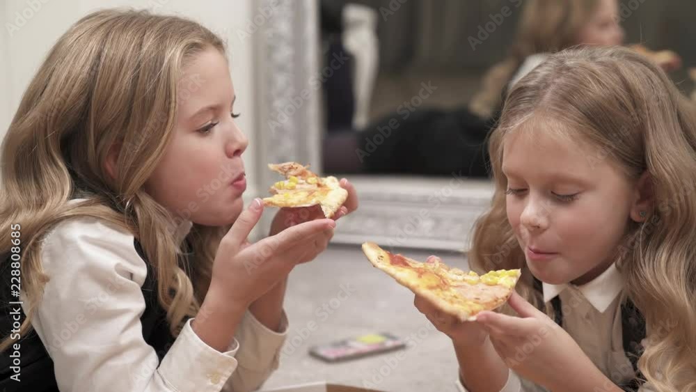 A close up of two charming girls lying on the floor and eating pizza. They face each other and talk while eating.
