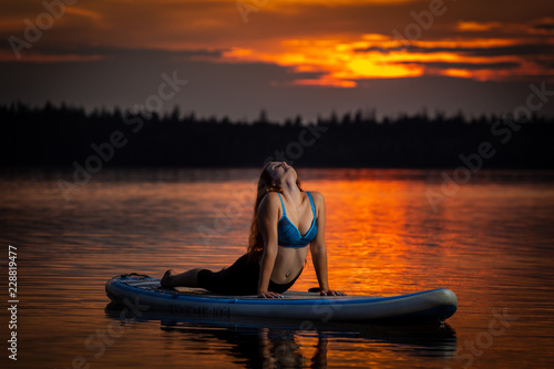 Beautiful slim fit girl with long brown hair exercising yoga on paddleboard in the dark, colorful sunset on scenic lake Velke Darko near Zdar nad Sazovou, Czech Republic photo
