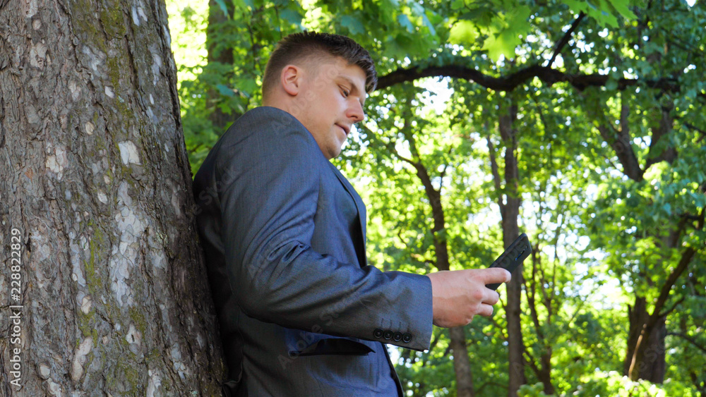 Young handsome guy (man) businessman standing by a tree in a suit. Concept: Nature, Rest, Suit, Businessman, Work, Lifestyle. Young handsome guy (man) businessman standing by a tree in a suit. Concept: Nature, Rest, Suit, Businessman, Work, Lifestyle.