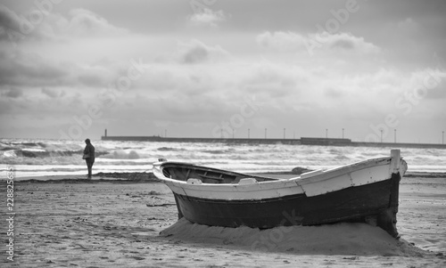 Moody black and white photo of an abandoned boat stuck in the sand on a beach with a single person in the distance near the shore with waves and clouds