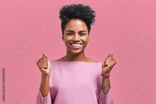 Portrait of overjoyed female clenches fists with with happiness, opens mouth widely as shouts loudly, celebrates her success, poses against pink background. People, happiness, success concept photo