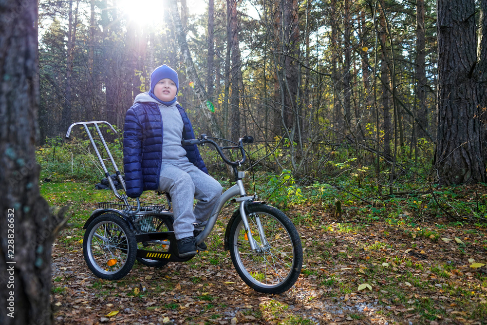 child autistic with disabilities on a tricycle with management for mom, in a blue cap and jacket in the Park for a walk