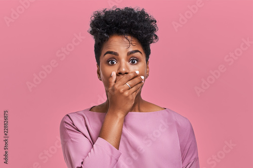 Horizontal shot of surprised African American woman covers mouth with both hands, tries to be speechless, looks with shocked expression, notices something strange, isolated over pink blank wall