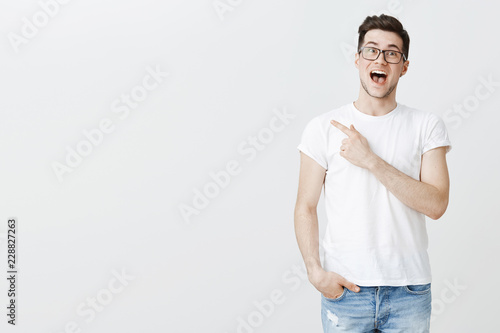 Portrait of very happy charming and attractive young man in glasses holding breath from excitement and amazement smiling with dropped jaw as pointing left at copy space being impressed and joyful
