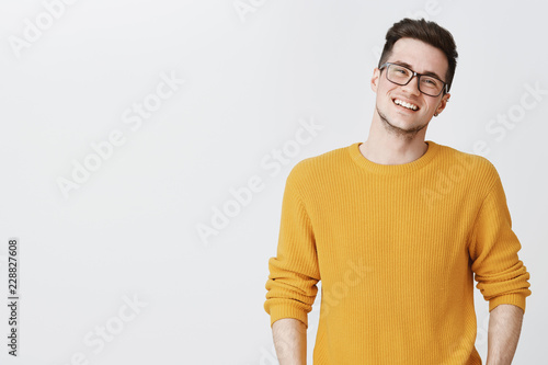Waist-up shot of happy and delighted handsome young man in glasses and yellow sweater tilting head, smiling and laughing as looking friendly at camera on right side of copy space over gray background photo