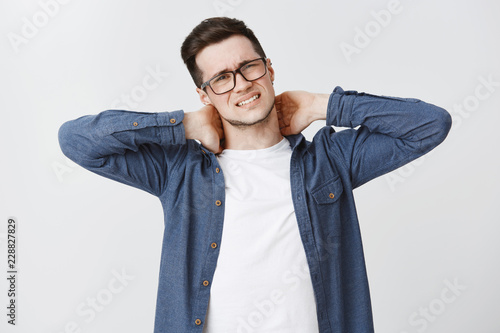 Waist-up shot of displeased young man feeling discomfort in neck touching back with hands frowning and clenching teeth from pain having problems with spine as sitting too long in front of computer