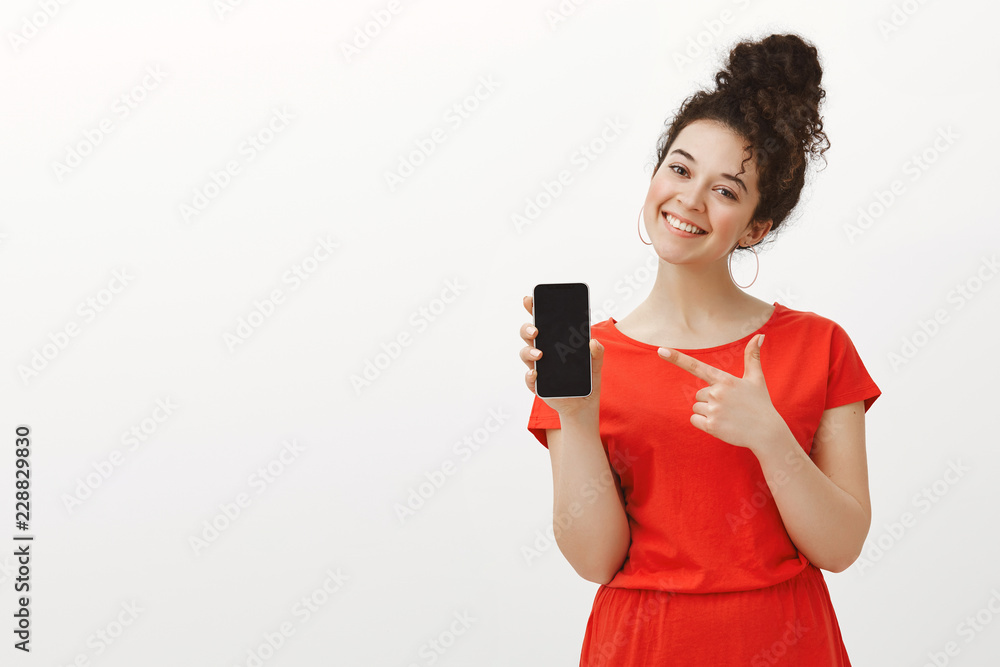 Pleased carefree girlfriend in cute red dress, showing smartphone and smiling broadly with tilted head while pointing at device with index finger, bragging about new gadger in front of girlfriends Pleased carefree girlfriend in cute red dress, showing smartphone and smiling broadly with tilted head while pointing at device with index finger, bragging about new gadger in front of girlfriends