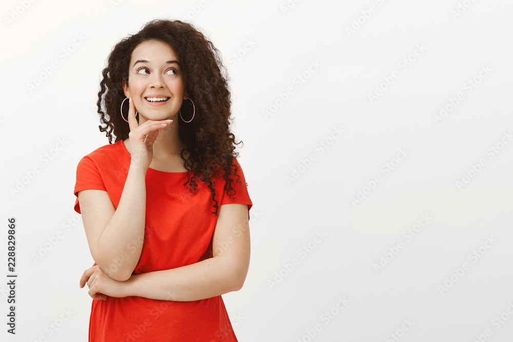 Portrait of dreamy good-looking stylish girlfriend with curly hair in red dress, smiling joyfully while looking aside, holding hand on chin, dreaming or imaging curious things, thinking over gray wall Portrait of dreamy good-looking stylish girlfriend with curly hair in red dress, smiling joyfully while looking aside, holding hand on chin, dreaming or imaging curious things, thinking over gray wall