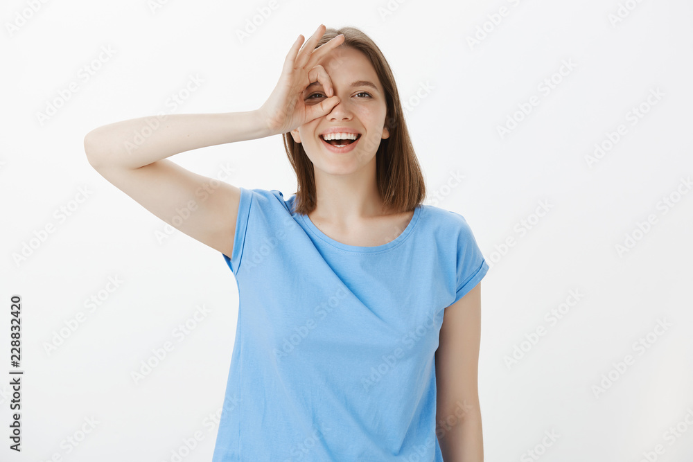 Optimistic girl looking forward to future adventures. Portrait of happy good-looking european woman in blue t-shirt, showing ok or okay gesture over eye and smiling broadly, having great time, playing Optimistic girl looking forward to future adventures. Portrait of happy good-looking european woman in blue t-shirt, showing ok or okay gesture over eye and smiling broadly, having great time, playing