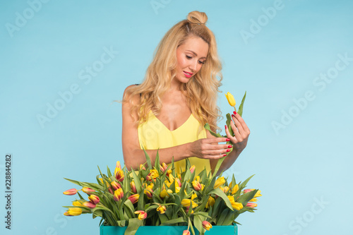 Floristics, holidays, flower shop and people concept - Beautiful blond young woman fixing bouquet of tulips on blue background.