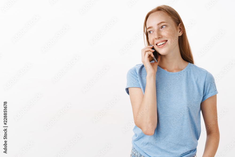Waist-up shot of carefree relaxed ordinary redhead european woman with freckles in blue t-shirt turning left while holding phone near ear and talking calling friend via smartphone to invite for dinner