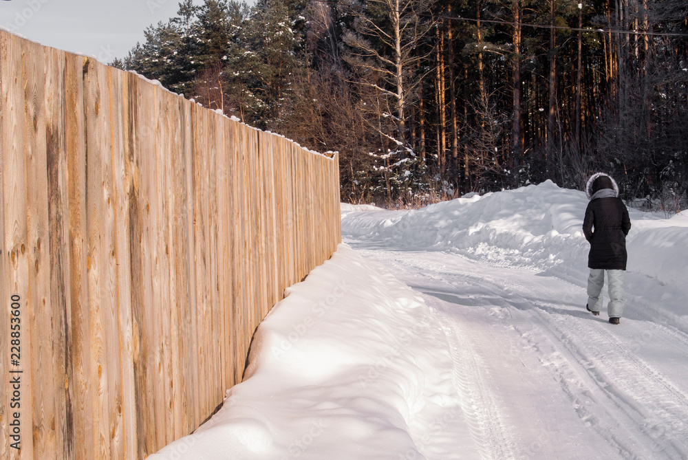 Obraz premium Girl walking on a snowy road along the wooden fence and forest in countryside