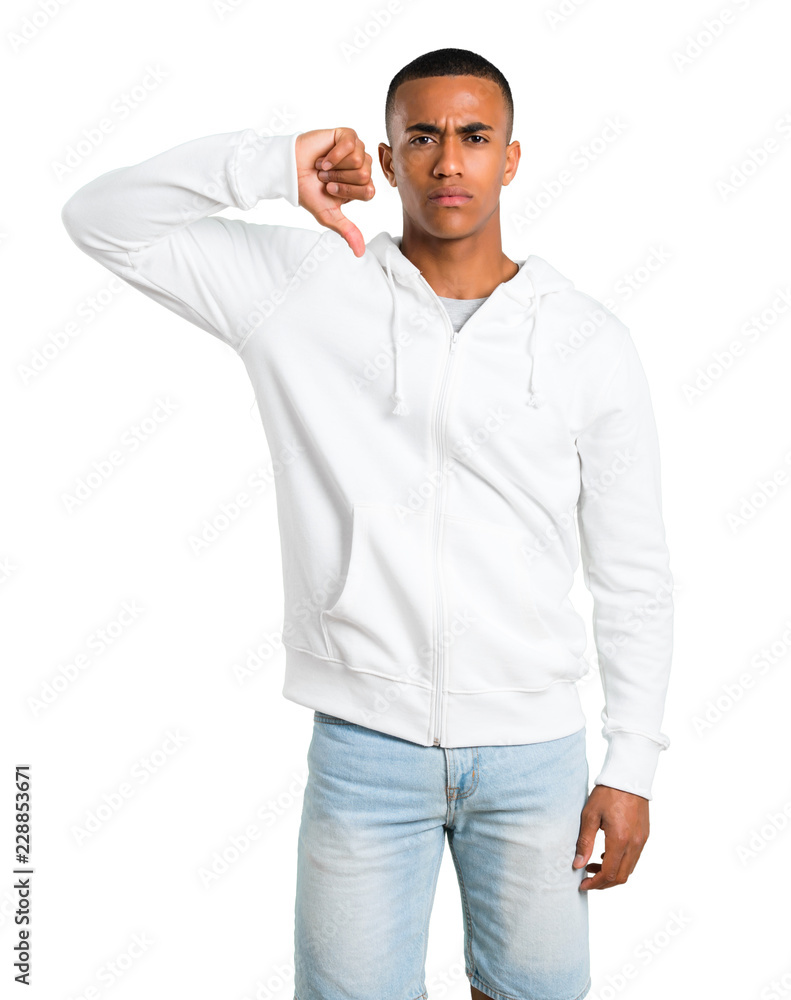 Dark-skinned young man with white sweatshirt showing thumb down sign with negative expression. Sad expression on isolated white background Dark-skinned young man with white sweatshirt showing thumb down sign with negative expression. Sad expression on isolated white background