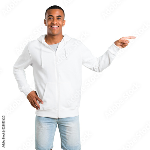 Dark-skinned young man with white sweatshirt pointing finger to the side and presenting a product while smiling in a confident pose on isolated white background