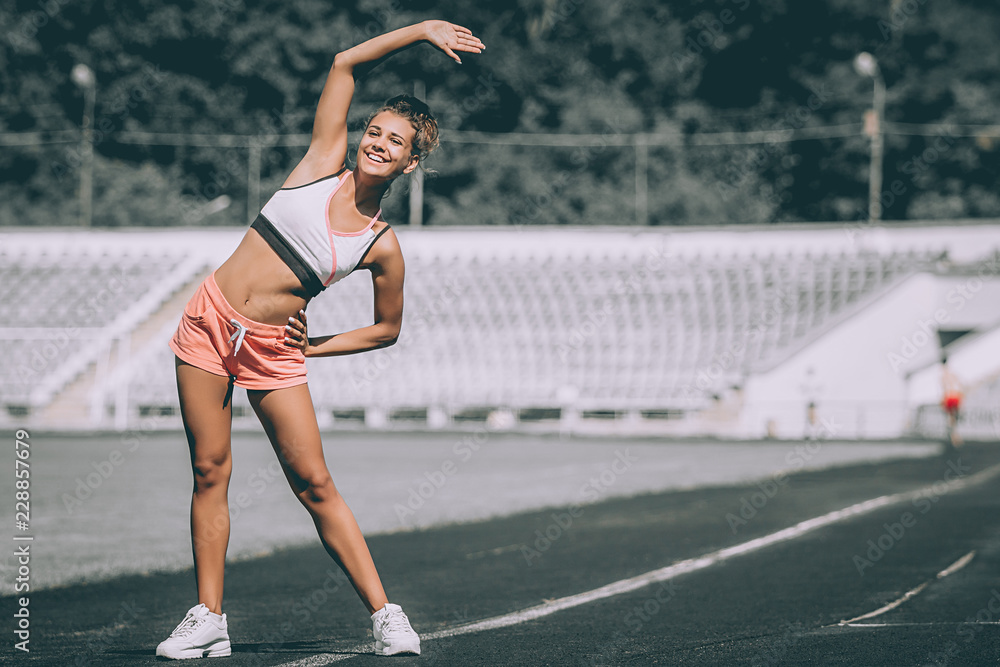 Sport, exercises outdoors. Girl doing stretching on stadium. Bending aside. Full body, looking aside. Grey background