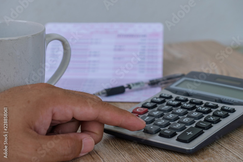 The finger is pressing the calculator on the wooden floor. Have a cup of coffee and bookbank next to it. photo