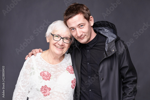  Family portrait of mature wrinkled gray haired woman dressed in stylish white blouse and son  who came to congratulate her with anniversary, have good relationships. Multi generation concept.