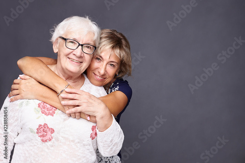  Family portrait of mature wrinkled gray haired woman dressed in stylish white blouse and daughter who came to congratulate her with anniversary, have good relationships. Multi generation concept.