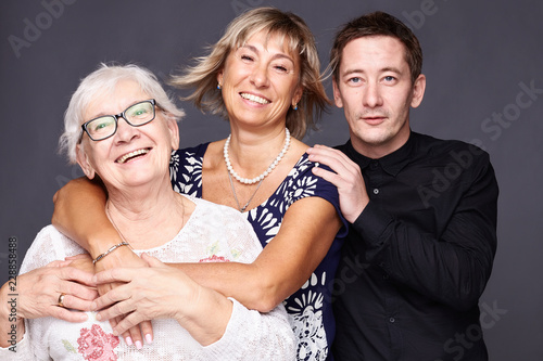 Multi generation concept. Family portrait of mature wrinkled woman dressed in stylish shirt, surrounded with love of  daughter and son who came to congratulate pretty elderly lady with anniversary.
