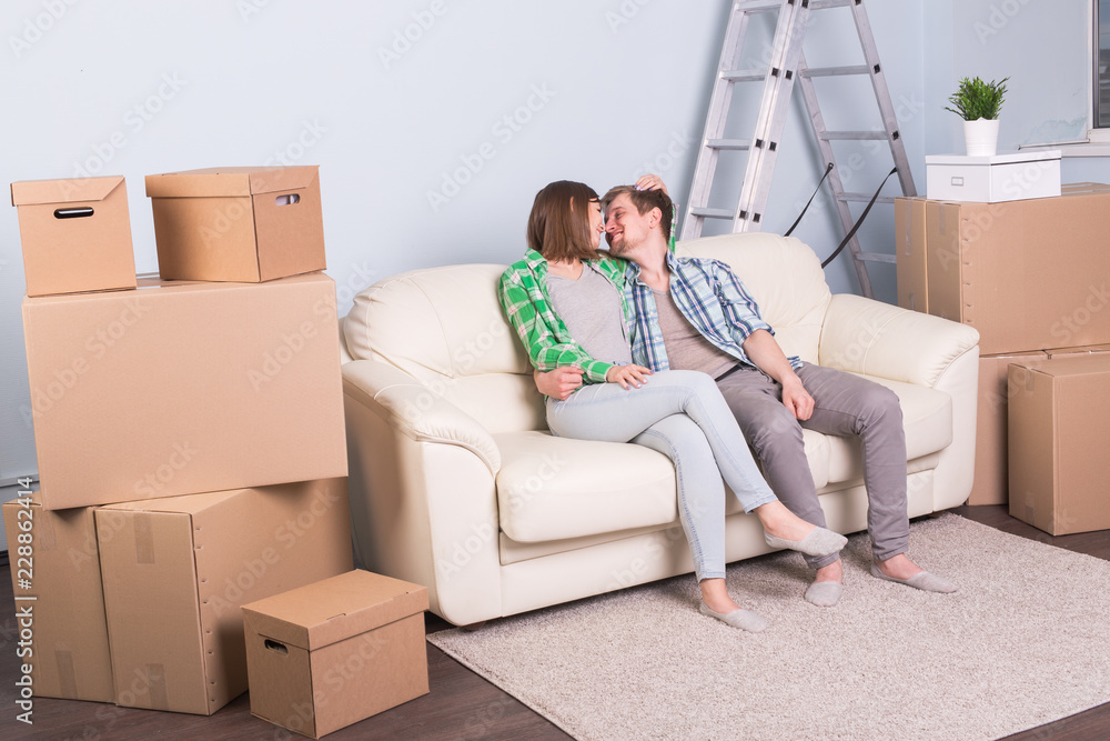 New home, relocation and moving concept - young couple sitting and kissing on the couch surrounded with boxes New home, relocation and moving concept - young couple sitting and kissing on the couch surrounded with boxes