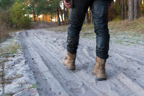A man in boots and leather pants is walking along a sandy road