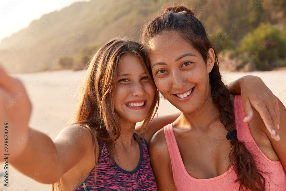 Mixed race women have good relations, pose for making selfie, going to share photos in social networks, make video call, pose against sandy beach. Joyful sisters spend summer vacation in exotic place Mixed race women have good relations, pose for making selfie, going to share photos in social networks, make video call, pose against sandy beach. Joyful sisters spend summer vacation in exotic place