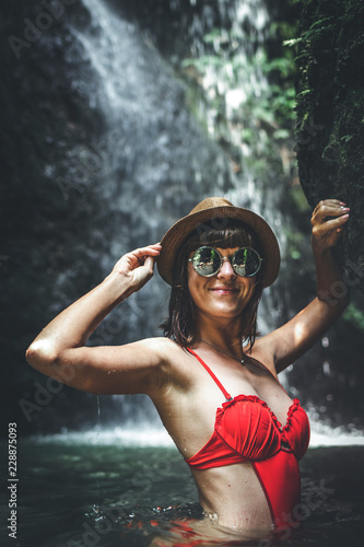 Fototapeta Naklejka Na Ścianę i Meble -  Young woman tourist in red swimsuit and straw hat posing in the deep jungle with waterfall. Real adventure concept. Bali island.