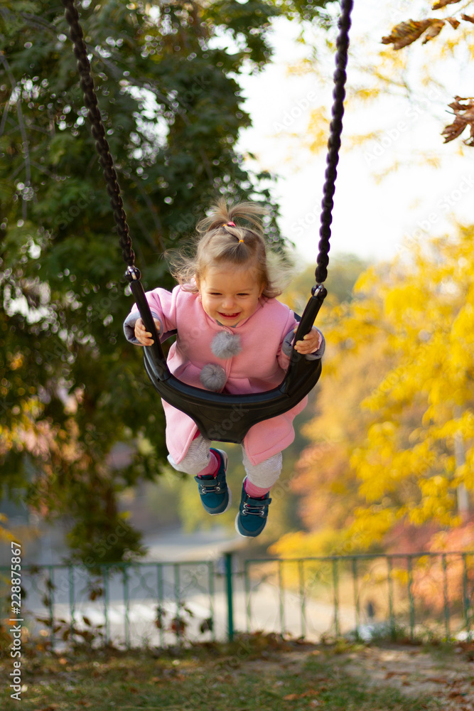Portrait of a joyful child riding on a swing in autumn park. Smiling child in a pink autumn coat while riding on a swing. Sweet little girl with curls and smiling eyes.