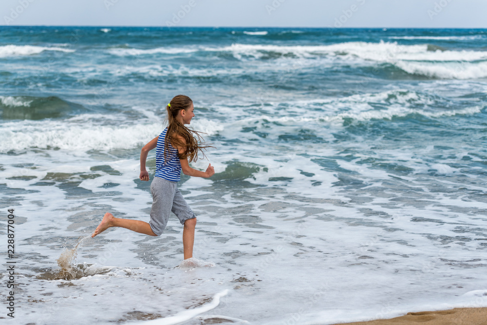 Cute happy little girl running along the beach in jumping over waves. Beautiful summer sunny day, blue sea, picturesque landscape.