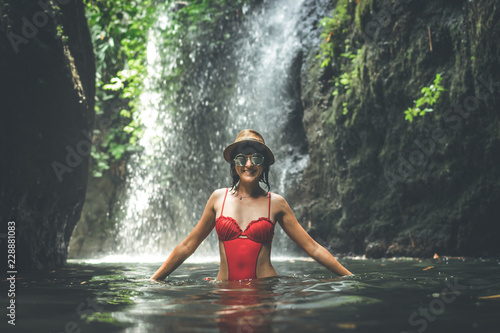 Fototapeta Naklejka Na Ścianę i Meble -  Young woman tourist with straw hat and red swimsuit in the deep jungle with waterfall. Real adventure concept. Bali island.