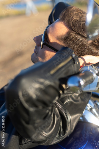 Young man wearing a black leather jacket, sunglasses and jeans lays outdoor on a motorcycle, on a mountain on a blue sky background. Lifestyle, travel.