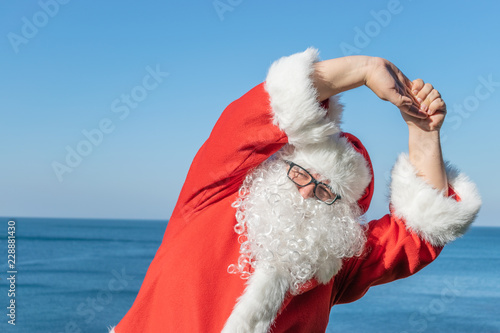 Santa doing exercises on the ocean. Traditional red outfit and relaxing on the beach