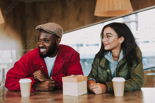 Cheerful relaxed couple sitting in the comfortable cafe with gift box on the table and smiling while looking into the distance photo