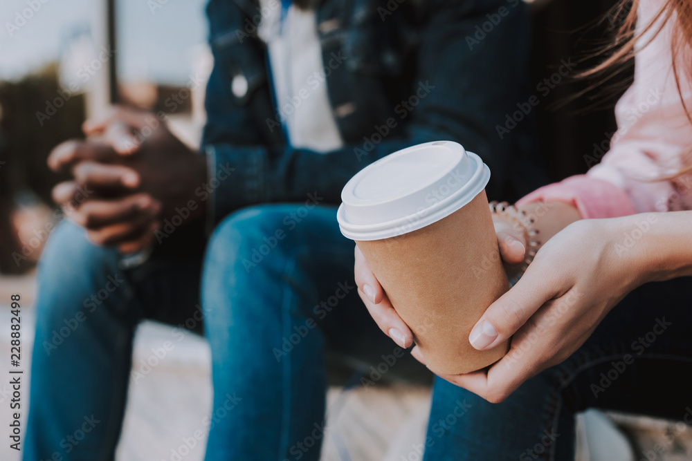 Close up of a cup with tasty coffee in female hands who is sitting with her Afro American friend