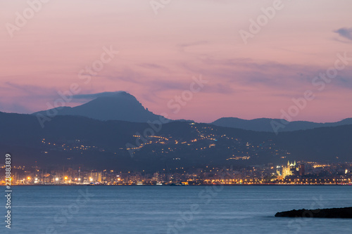 Colorful sunset at Mallorca island. Mediterranean sea and lights of Palma city with blue mountains Serra de Tramuntana and pink evening sky in background.