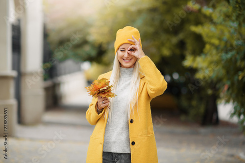 Outdoors lifestyle fashion portrait of happy pretty girl walking on the autumn park. Holding maple leaves. Wearing in yellow coat and hat.  Smiling  enjoying autumn nature. Jogging  having fun