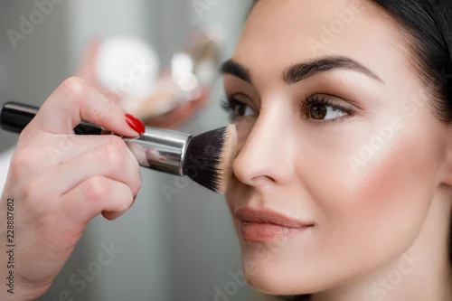 Fototapeta Close up of makeup artist applying light layer of matting powder while using professional brush for lady