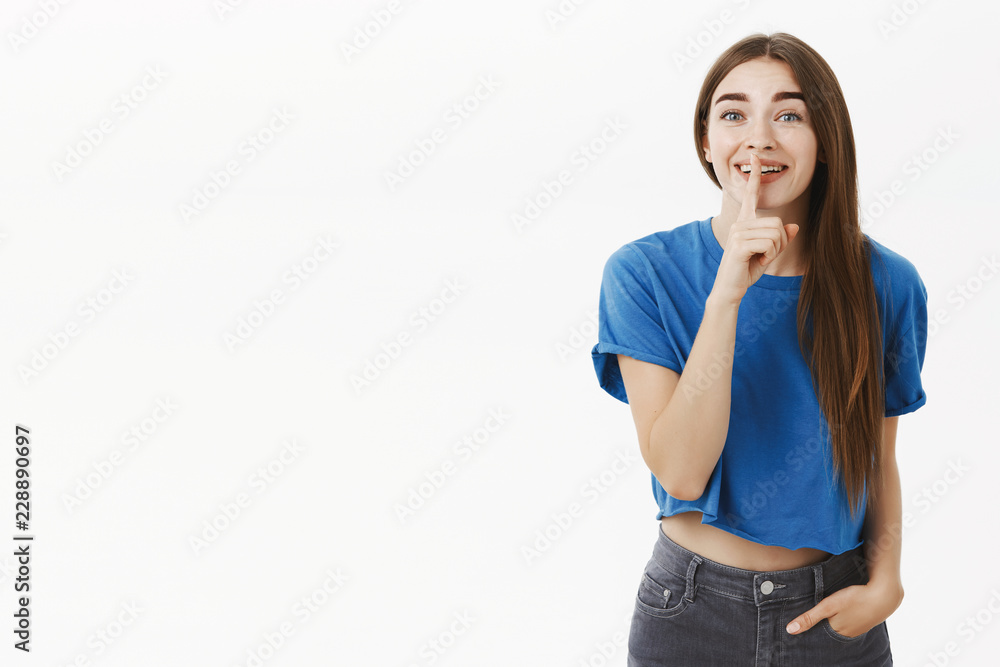 Woman talking with friend chuckling saying shh at camera asking keep quiet so no one listen funny rumor, gossiping making shush gesture with index finger over mouth, smiling joyfully over grey wall Woman talking with friend chuckling saying shh at camera asking keep quiet so no one listen funny rumor, gossiping making shush gesture with index finger over mouth, smiling joyfully over grey wall