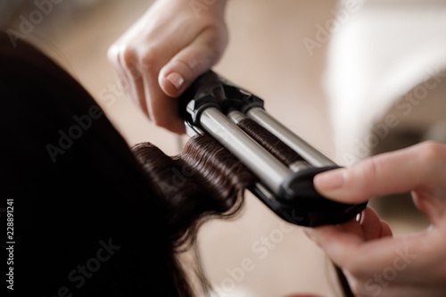 Close-up of stylist hands holding frizzling iron. Master is waving long straight strands of woman using modern tools