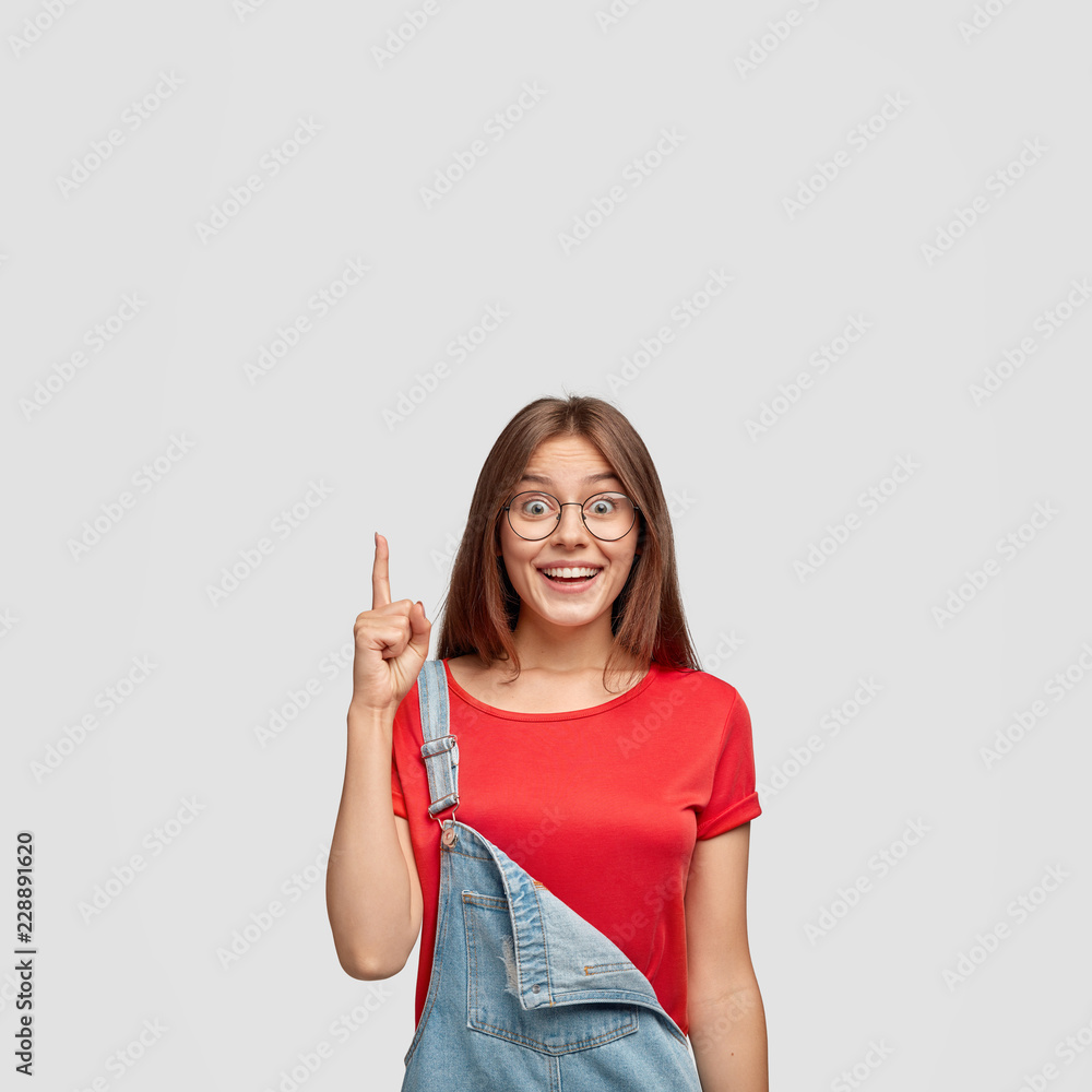 Indoor shot of good looking satisfied brunette woman has pleased facial expression, gets brilliant idea in mind, points with fore finger upwards, dressed casually, isolated over white background Indoor shot of good looking satisfied brunette woman has pleased facial expression, gets brilliant idea in mind, points with fore finger upwards, dressed casually, isolated over white background