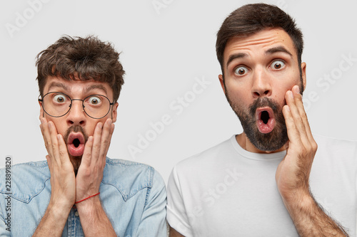 Shocked two guys stares with bugged eyes at camera, keep hands on cheeks, wonder about latest news, pose against white background. Emotive Caucasian men express great surprisement or disbelief