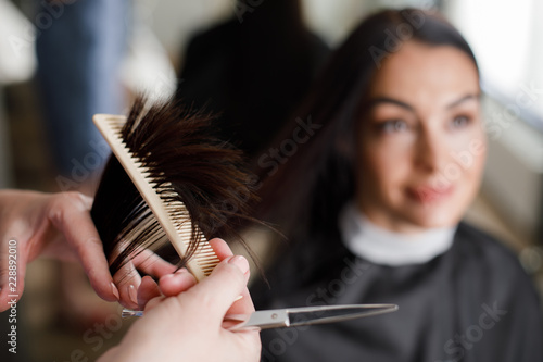 Focus on close-up of stylist hands combing female hair. She is holding scissors for cutting split ends while smiling customer is sitting in chair