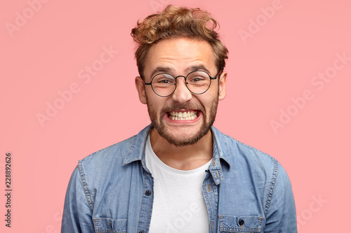 Photo of pleased curly Caucasian man smiles joyfully at camera, clenches teeth, dressed in fashionable blue shirt, being in good mood after date with girlfriend, poses against pink background