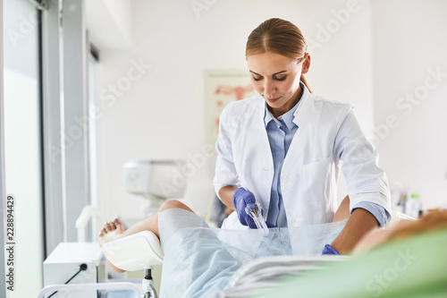 Waist up portrait of gynecologist in white lab coat and sterile gloves using vaginal speculum during pelvic exam. She is looking at instrument and smiling