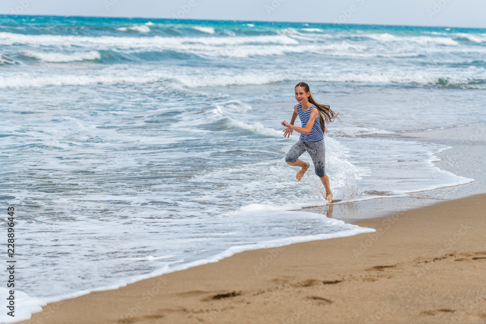 Cute happy Girl in striped t-shirt running along the beach in jumping over waves. Beautiful summer sunny day, blue sea, picturesque landscape. Cute happy Girl in striped t-shirt running along the beach in jumping over waves. Beautiful summer sunny day, blue sea, picturesque landscape.